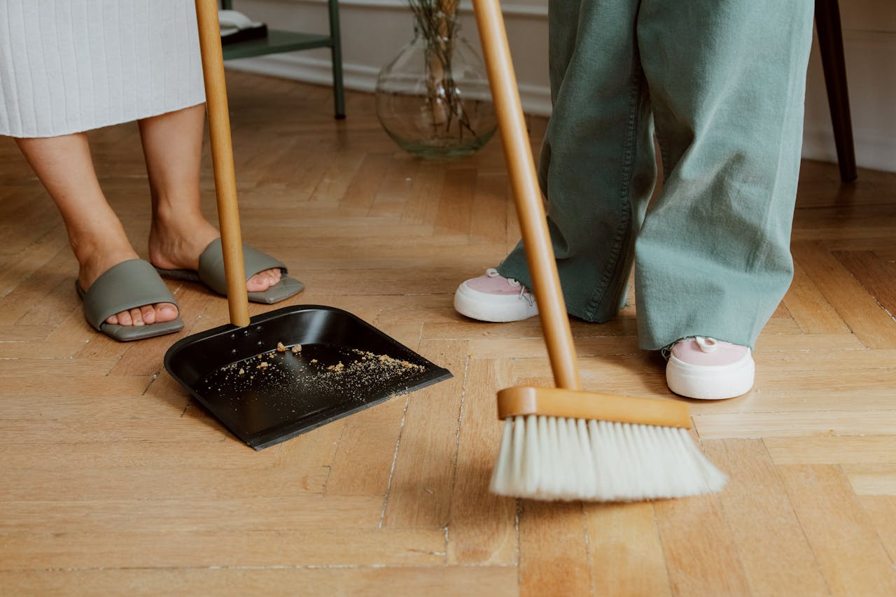 Close-up of people cleaning wooden floor with broom and dustpan indoors.