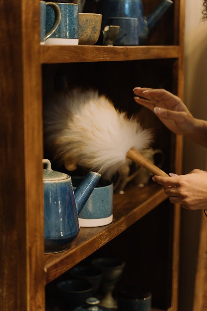 Hands cleaning pottery on a wooden shelf with a dusting brush, emphasizing home maintenance.